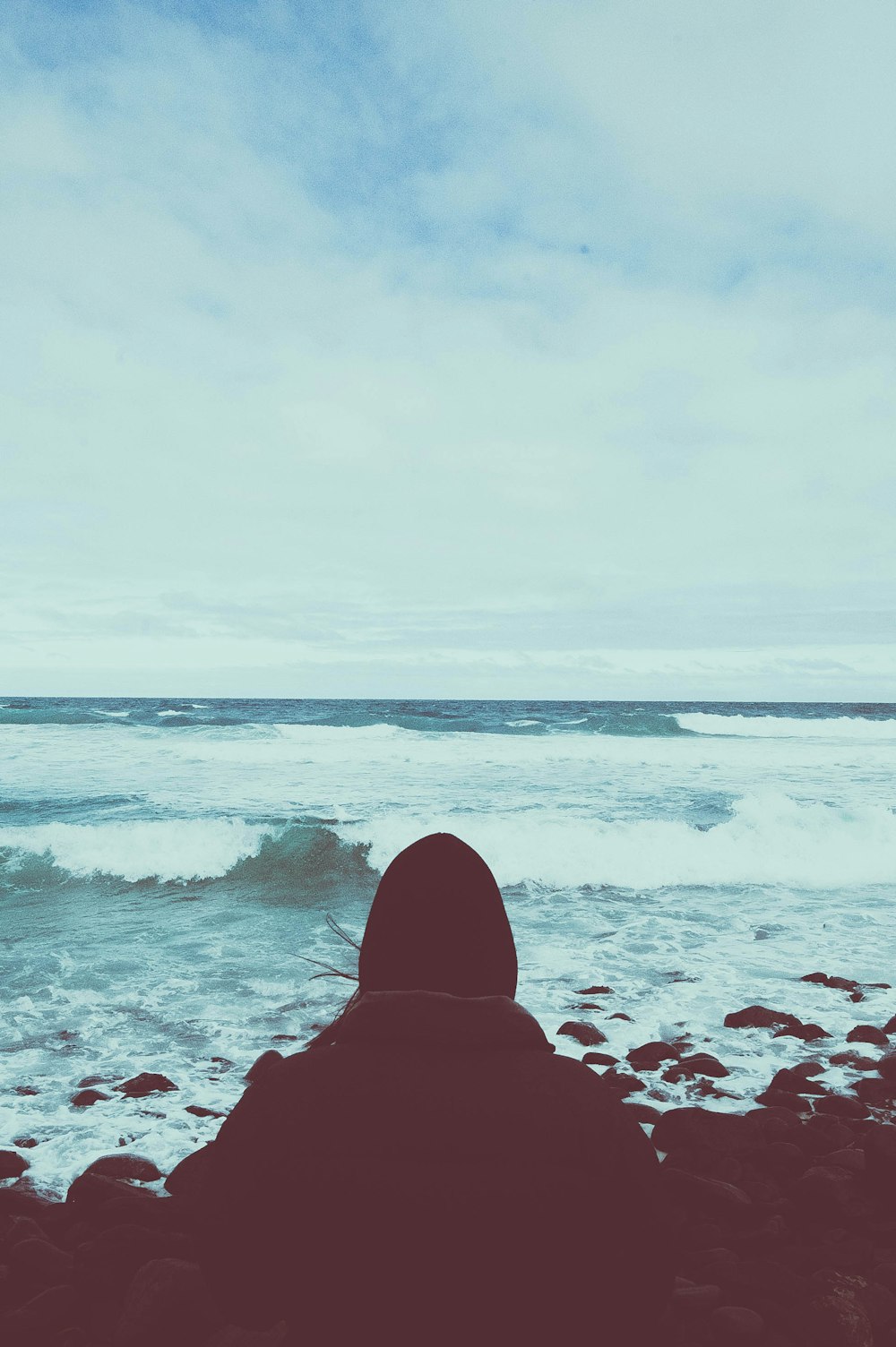 person sitting on rocking shore under cloudy sky