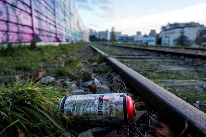 A spray paint can lies on the ground beside a railway track, surrounded by grass and gravel. The background features a long wall covered in colorful graffiti and distant city buildings.