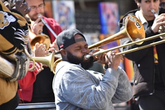 A group of musicians play brass instruments energetically. The focus is on a bearded man wearing a cap, who is playing the trombone with a passionate expression. Other musicians are seen in the background, contributing with trumpets, and they are dressed casually.