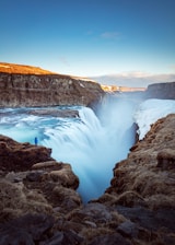 person standing on cliff near large waterfalls under blue sky at daytime Iceland Greenland Nordics