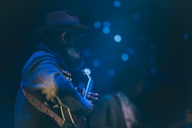 A person wearing a hat and thick coat is playing a guitar. The scene is dimly lit with a soft focus on the musician, creating a moody atmosphere. The background is filled with blurry blue lights that resemble bokeh, adding to the dreamlike quality of the image.