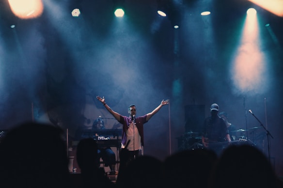 A singer with a microphone stands on stage with arms raised, surrounded by musicians playing various instruments. The stage is illuminated by spotlights and engulfed in a smoky atmosphere, suggesting a live music performance.