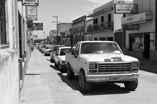 A black and white image of an urban street featuring several parked cars, including a prominently displayed old pickup truck with a weathered exterior. The street is lined with various buildings that have visible signs with text in Spanish. Overhead power lines and a clear sky are in the background.