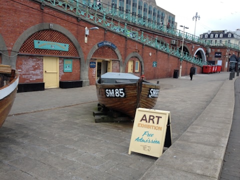 A wooden boat with the markings SM85 is placed on a concrete pathway in front of arched brick structures. A sign next to the boat advertises an art exhibition with free admission. The background has red brick walls with teal signage and a series of archways. Some people can be seen walking along the path.