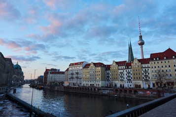A scenic urban landscape featuring a river running alongside a row of historic buildings with pastel-colored facades and red roofs. In the background, a tall television tower and a cathedral with a green dome are visible under a blue sky with pinkish clouds.