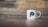 A white enamel mug with black trim sits on a wooden surface. The mug features a black design with the text 'DECAF AND CO' inside a stylized skull graphic. The background is a blurred wooden plank wall, adding a rustic and minimalist atmosphere.