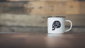 A white enamel mug with black trim sits on a wooden surface. The mug features a black design with the text 'DECAF AND CO' inside a stylized skull graphic. The background is a blurred wooden plank wall, adding a rustic and minimalist atmosphere.