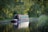 A narrowboat is gently floating on a calm canal, surrounded by lush green foliage. The sunlight filters through the trees, casting dappled light onto the water surface and creating a peaceful scene. The boat's reflection can be seen in the tranquil water.