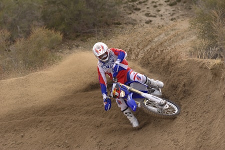 A motocross rider in full gear is skillfully maneuvering a dirt bike on a sandy track, creating a cloud of dust as the bike leans into a turn. The rider is wearing red, white, and blue gear, with a helmet that matches the color scheme. Surrounding the track are dry bushes and foliage, indicating an outdoor, natural setting.