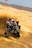 A person is riding an all-terrain vehicle (ATV) aggressively over sand dunes. The vehicle is kicking up a large cloud of sand as it moves, and a red flag is attached to it. The background is a vast desert landscape.