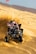A person is riding an all-terrain vehicle (ATV) aggressively over sand dunes. The vehicle is kicking up a large cloud of sand as it moves, and a red flag is attached to it. The background is a vast desert landscape.