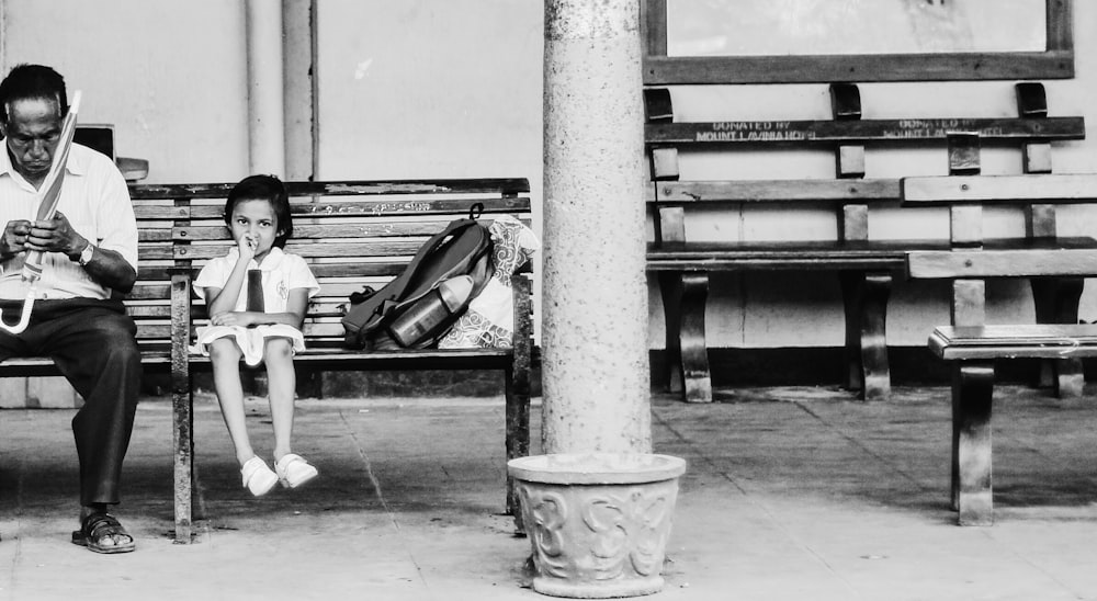 Fotografía en escala de grises de hombre sosteniendo paraguas sentado en un banco al lado de la niña