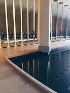 A series of tall, vertical mirrors are positioned in an indoor space, reflecting a group of people standing nearby. The floor is made of light-colored tiles, and there is a dark, reflective pool of water adjacent to the mirrors, creating additional reflections.