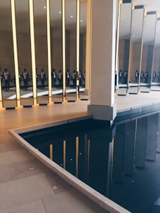 A series of tall, vertical mirrors are positioned in an indoor space, reflecting a group of people standing nearby. The floor is made of light-colored tiles, and there is a dark, reflective pool of water adjacent to the mirrors, creating additional reflections.