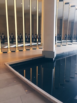 A series of tall, vertical mirrors are positioned in an indoor space, reflecting a group of people standing nearby. The floor is made of light-colored tiles, and there is a dark, reflective pool of water adjacent to the mirrors, creating additional reflections.