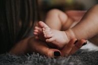 Infant's feet being held by a woman's hand with painted and manicured hands resting on a gray blanket