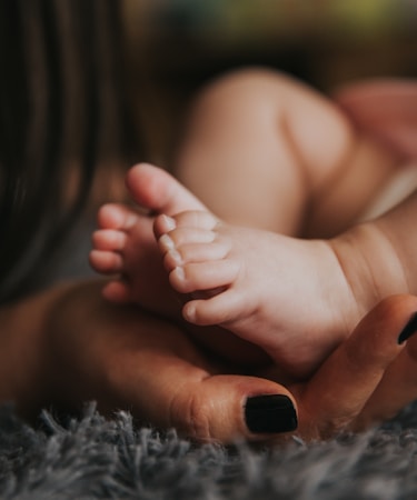 Infant's feet being held by a woman's hand with painted and manicured hands resting on a gray blanket