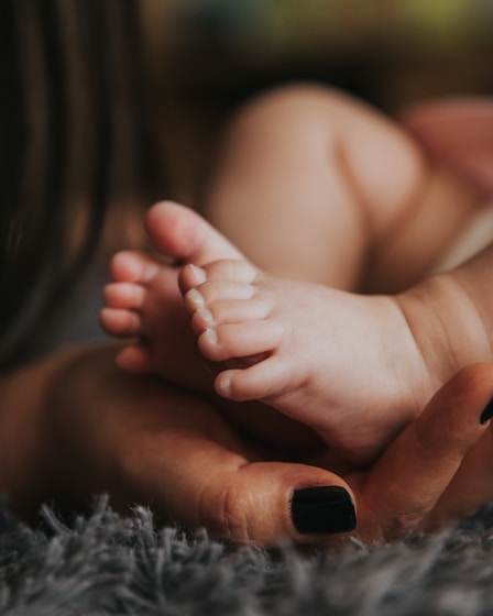 Infant's feet being held by a woman's hand with painted and manicured hands resting on a gray blanket