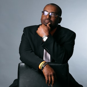 A person in formal attire sits pensively, resting a hand on their chin. They wear glasses, a black suit with an orange shirt, and a patterned tie. A watch and bracelets adorn one wrist, and they are seated on a black leather chair against a plain background.