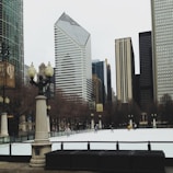 An outdoor ice skating rink surrounded by tall buildings in an urban area. The architecture includes modern skyscrapers and classical-style columns with ornamental street lamps. Several people are skating on the ice, adding a lively atmosphere.
