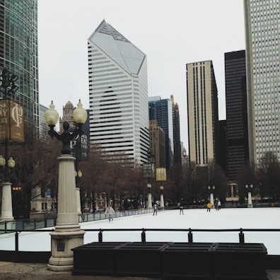 An outdoor ice skating rink surrounded by tall buildings in an urban area. The architecture includes modern skyscrapers and classical-style columns with ornamental street lamps. Several people are skating on the ice, adding a lively atmosphere.