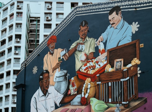 A vibrant mural depicts four individuals engaged in various traditional activities. One person, dressed in a traditional outfit with a red turban, is pouring a liquid into a cup. Another individual is holding a glass jar, while a man in light clothing is stringing together garlands of flowers. The fourth person appears to be attending to a birdcage that has several garlands and a photo resting on top. A green parrot is perched near the cage, adding more color and cultural significance to the scene. The backdrop features a tall residential building, giving the mural an urban setting.
