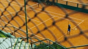 Viewed through a chain-link fence, a tennis player on a clay court is preparing to serve. The scene includes well-marked tennis courts, concrete steps, and green vegetation, creating a dynamic perspective.