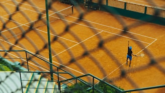 Viewed through a chain-link fence, a tennis player on a clay court is preparing to serve. The scene includes well-marked tennis courts, concrete steps, and green vegetation, creating a dynamic perspective.