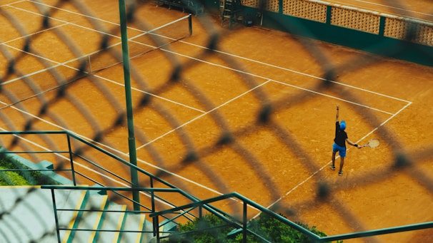 Viewed through a chain-link fence, a tennis player on a clay court is preparing to serve. The scene includes well-marked tennis courts, concrete steps, and green vegetation, creating a dynamic perspective.