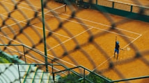 Viewed through a chain-link fence, a tennis player on a clay court is preparing to serve. The scene includes well-marked tennis courts, concrete steps, and green vegetation, creating a dynamic perspective.