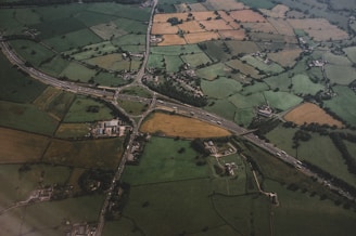 An aerial view of a rural landscape featuring a patchwork of agricultural fields in various shades of green, brown, and yellow. A network of roads and a highway interchange cuts through the countryside, connecting scattered buildings and small clusters of houses. The terrain is slightly rolling with fields divided by hedgerows and trees.