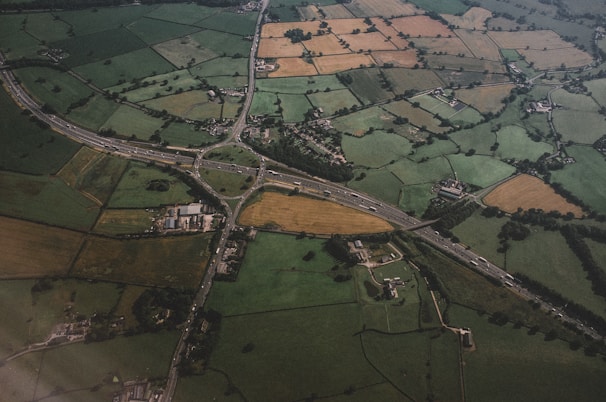 An aerial view of a rural landscape featuring a patchwork of agricultural fields in various shades of green, brown, and yellow. A network of roads and a highway interchange cuts through the countryside, connecting scattered buildings and small clusters of houses. The terrain is slightly rolling with fields divided by hedgerows and trees.