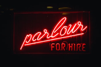 A bright red neon sign with the words 'parlour' written in elegant cursive and 'FOR HIRE' in block letters underneath. The background is dark, making the vividness of the neon light stand out prominently.