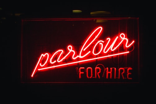 A bright red neon sign with the words 'parlour' written in elegant cursive and 'FOR HIRE' in block letters underneath. The background is dark, making the vividness of the neon light stand out prominently.
