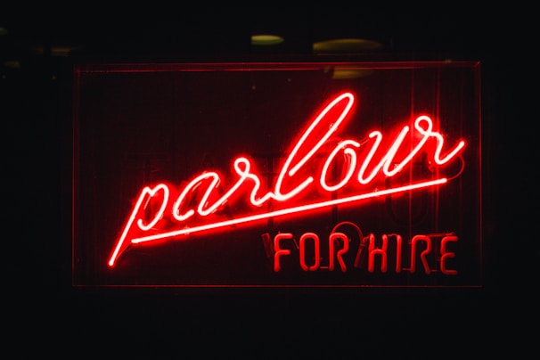 A bright red neon sign with the words 'parlour' written in elegant cursive and 'FOR HIRE' in block letters underneath. The background is dark, making the vividness of the neon light stand out prominently.