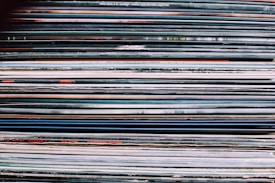 A stack of vinyl record covers is tightly packed together, showing a variety of colors and text on their spines. The spines display different artists' names and album titles, with a mix of bold and thin typography. The overall texture suggests the collection is aged or well-used.