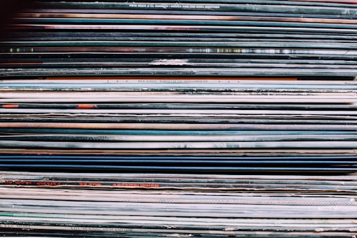 A stack of vinyl record covers is tightly packed together, showing a variety of colors and text on their spines. The spines display different artists' names and album titles, with a mix of bold and thin typography. The overall texture suggests the collection is aged or well-used.