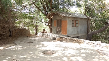 A small, rustic house with a thatched roof stands surrounded by dense trees, creating a secluded, peaceful atmosphere. The house is made of rough materials with wooden shutters on the windows. The area around the house is paved with stones, leading up to a circular stone structure in the foreground.
