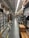 An aisle in a hardware store lined with shelves stocked with various products, including boxes, cans, and construction materials. The aisle is long and narrow, flanked by high shelves filled with merchandise. The ceiling features exposed utilities and fluorescent lighting.