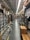 An aisle in a hardware store lined with shelves stocked with various products, including boxes, cans, and construction materials. The aisle is long and narrow, flanked by high shelves filled with merchandise. The ceiling features exposed utilities and fluorescent lighting.