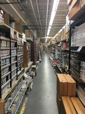 An aisle in a hardware store lined with shelves stocked with various products, including boxes, cans, and construction materials. The aisle is long and narrow, flanked by high shelves filled with merchandise. The ceiling features exposed utilities and fluorescent lighting.