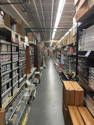 An aisle in a hardware store lined with shelves stocked with various products, including boxes, cans, and construction materials. The aisle is long and narrow, flanked by high shelves filled with merchandise. The ceiling features exposed utilities and fluorescent lighting.