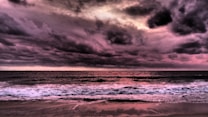 A dramatic seascape with deep, turbulent clouds casting shades of pink and purple over the ocean. The horizon is defined by the meeting of the churning sea and the vibrant sky. Waves with foamy crests gently wash onto the sandy shore, reflecting the colors of the sky.
