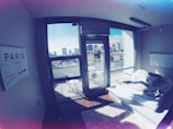 A modern apartment bedroom with large windows showcasing a city skyline. The room contains a simple platform bed with white bedding and fluffy rugs. A sign on the wall points towards Paris, and there are outdoor chairs on a small balcony.