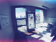 A modern apartment bedroom with large windows showcasing a city skyline. The room contains a simple platform bed with white bedding and fluffy rugs. A sign on the wall points towards Paris, and there are outdoor chairs on a small balcony.