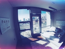 A modern apartment bedroom with large windows showcasing a city skyline. The room contains a simple platform bed with white bedding and fluffy rugs. A sign on the wall points towards Paris, and there are outdoor chairs on a small balcony.