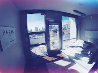 A modern apartment bedroom with large windows showcasing a city skyline. The room contains a simple platform bed with white bedding and fluffy rugs. A sign on the wall points towards Paris, and there are outdoor chairs on a small balcony.
