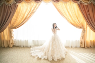 A bride wearing an elegant and ornate wedding dress stands gracefully before flowing golden drapes that are drawn back to allow sunlight to flood into the room. The scene is lavish and bathed in light, highlighting the intricate lace details of the gown.