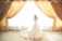 A bride wearing an elegant and ornate wedding dress stands gracefully before flowing golden drapes that are drawn back to allow sunlight to flood into the room. The scene is lavish and bathed in light, highlighting the intricate lace details of the gown.