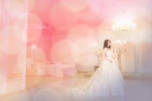 A woman wearing a long, elegant bridal gown in an opulent setting with soft, ambient lighting and a large chandelier. The room features decorative panels on the walls, a lavish candelabra, and a round table with chairs covered in white fabric creating a luxurious and dreamy atmosphere.